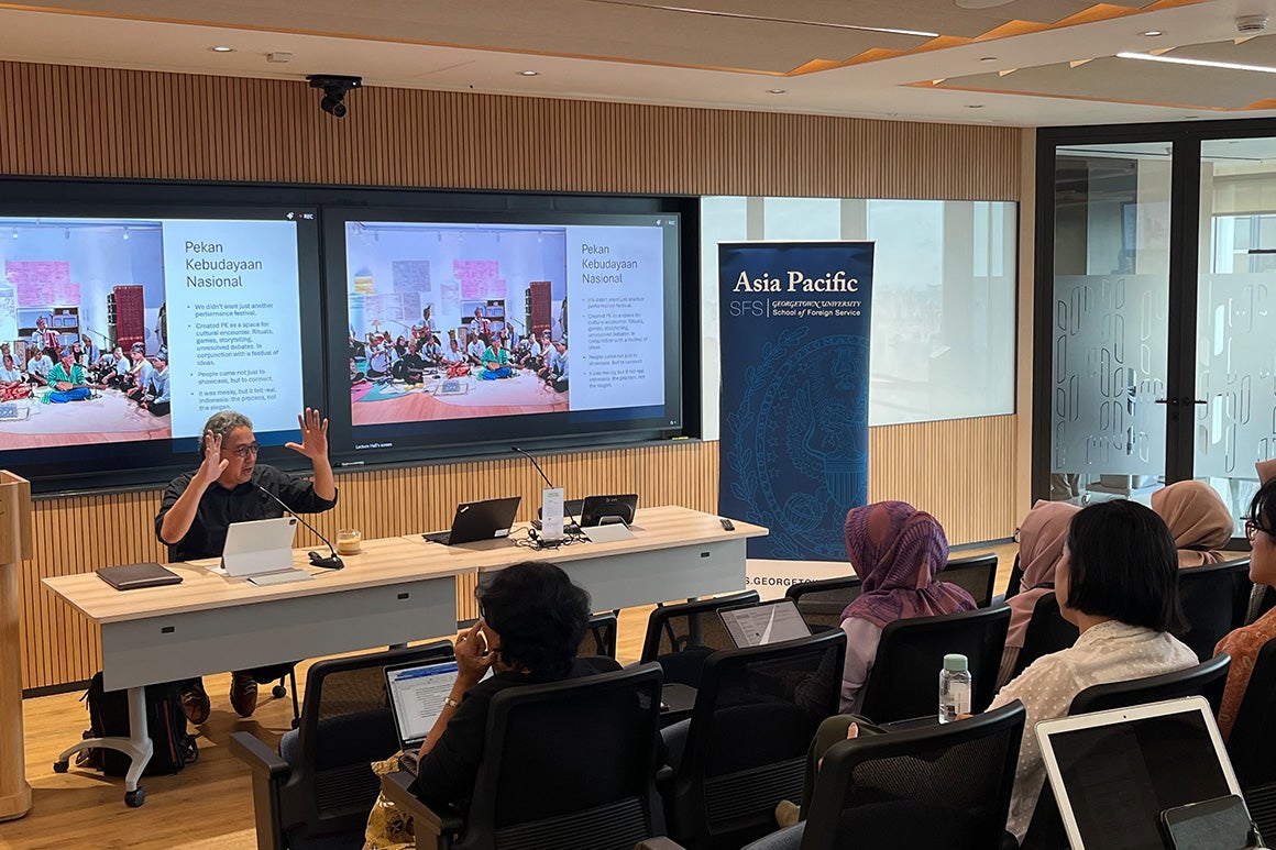 A professor speaking to students in a classroom with a presentation displayed behind them