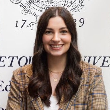 Headshot of Gemma King wearing a brown suit, standing in front of a Georgetown wall.