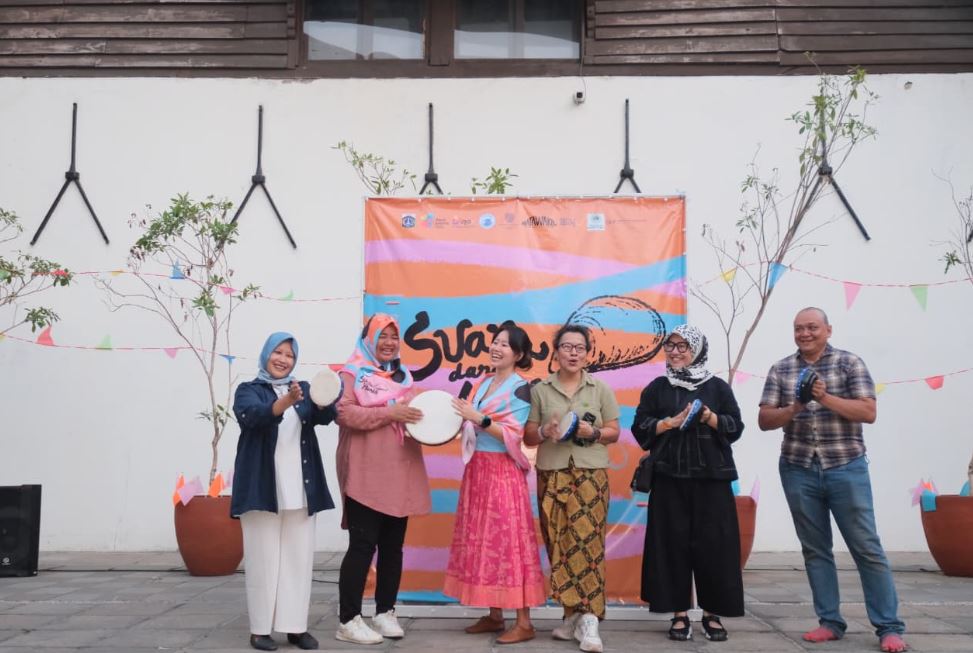 Six people stand in front of a 'Suara dari Muara' exhibition banner, smiling and holding tambourines during the exhibition opening.