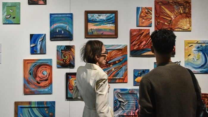 Two people stand in an art gallery viewing a wall filled with colorful abstract paintings.